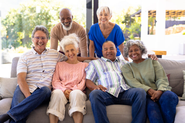 Portrait of happy multiracial male and female senior friends relaxing in living room at nursing home. Smiling, sofa, window, unaltered, togetherness, support, assisted living and retirement concept.